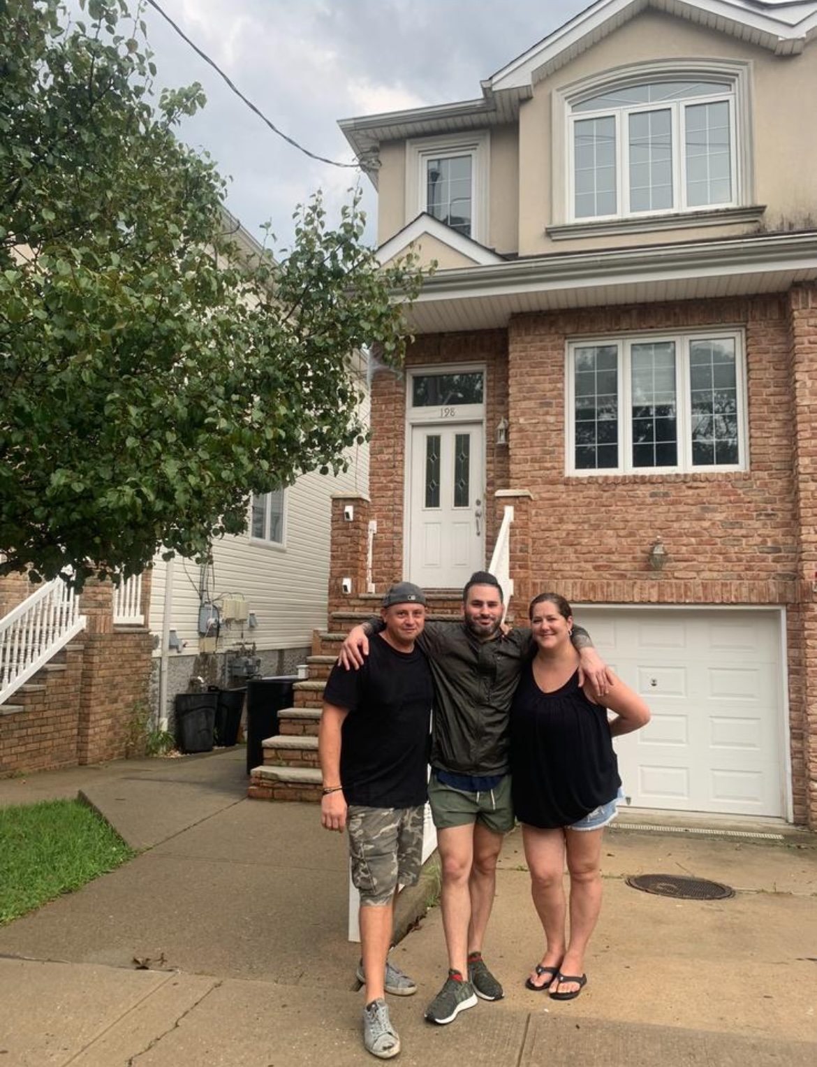 Three people standing outside in front of a brick house, arms around each other, smiling
