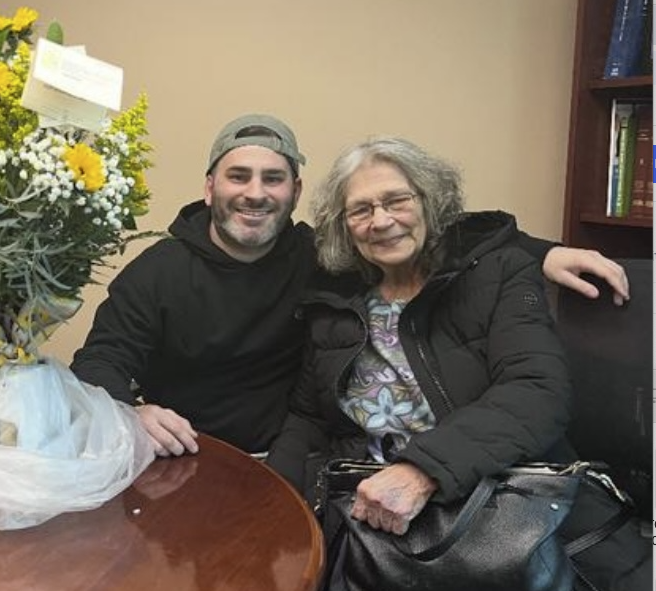 A man and older woman sitting together at a table with large bouquets of flowers and plants in front of them, smiling warmly